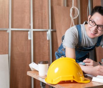 asian attractive glasses interior designer working at house site renovation construction progress with smile and confident at desk with blue print and drawing tool